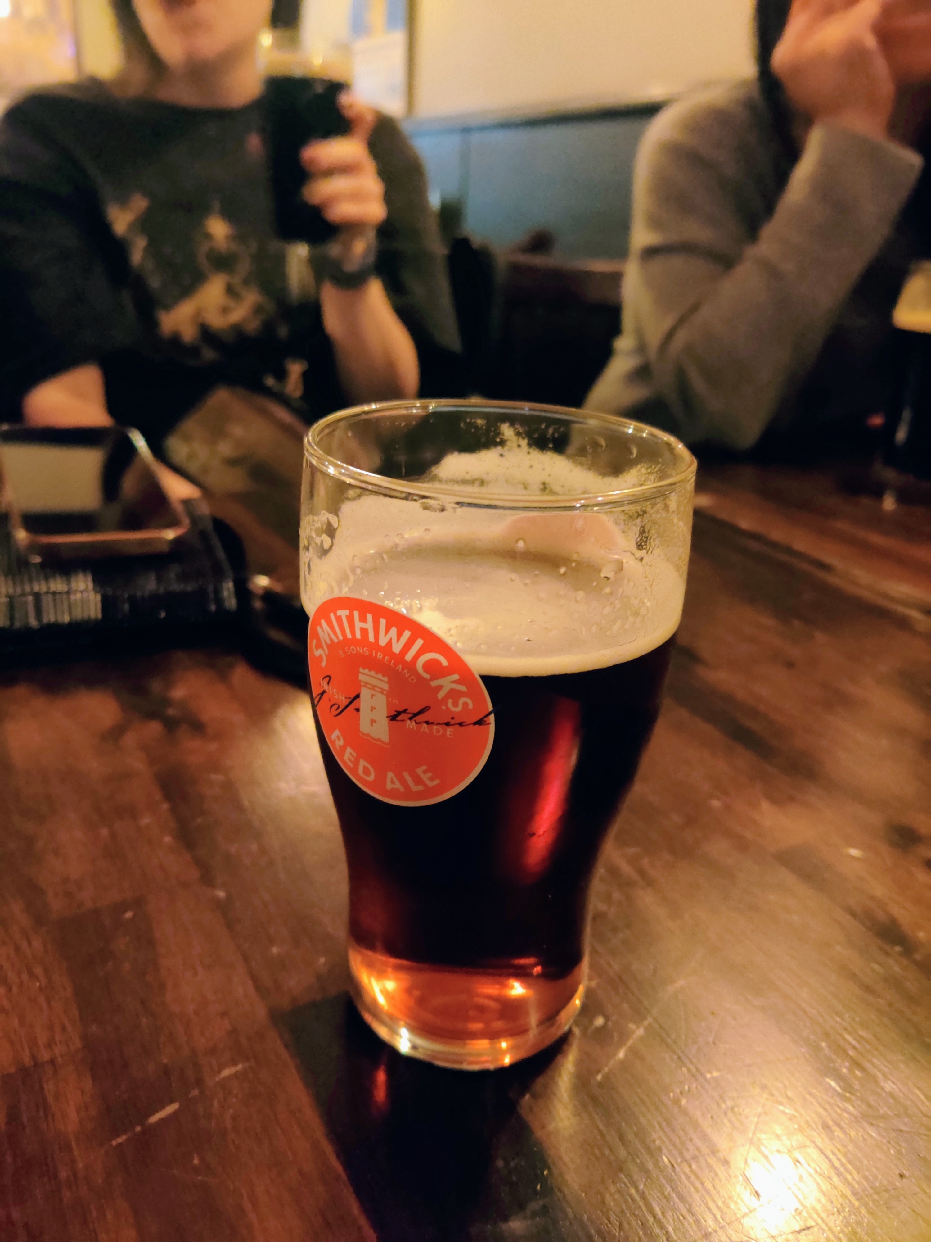 A pint of Smithwicks ale on a wooden table. Two ladies with their beers are in the background.