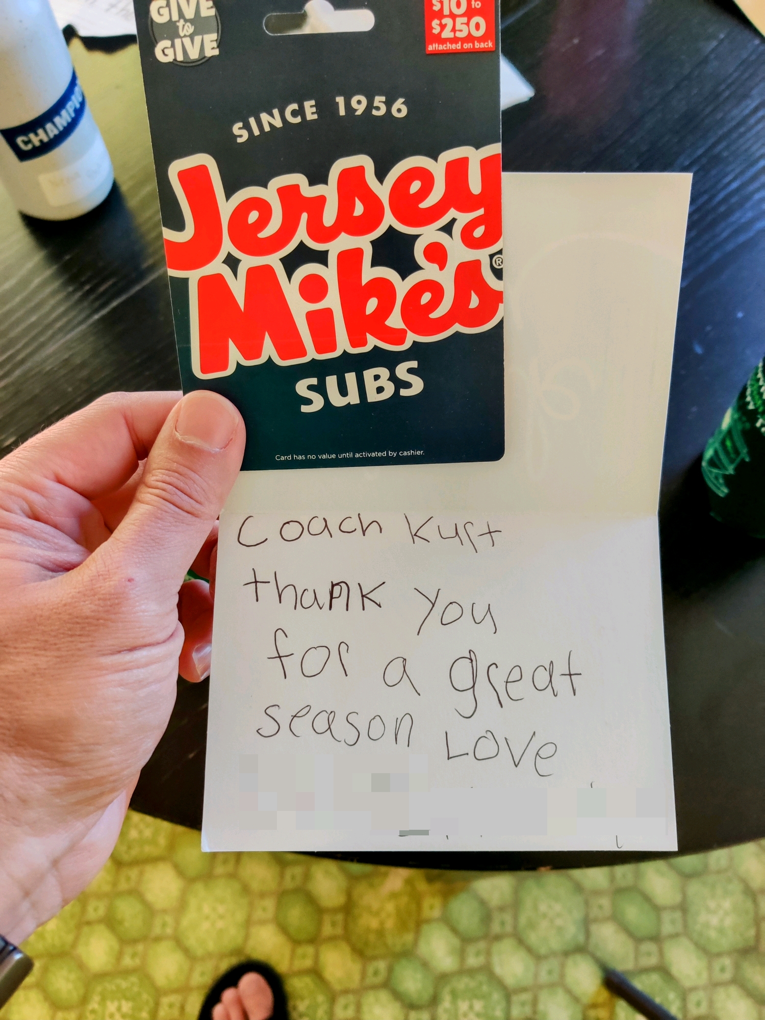 POV: you're holding a Jersey Mike's gift card and a thank you card with child's handwriting. A black kitchen table and green floor are visible in the background.