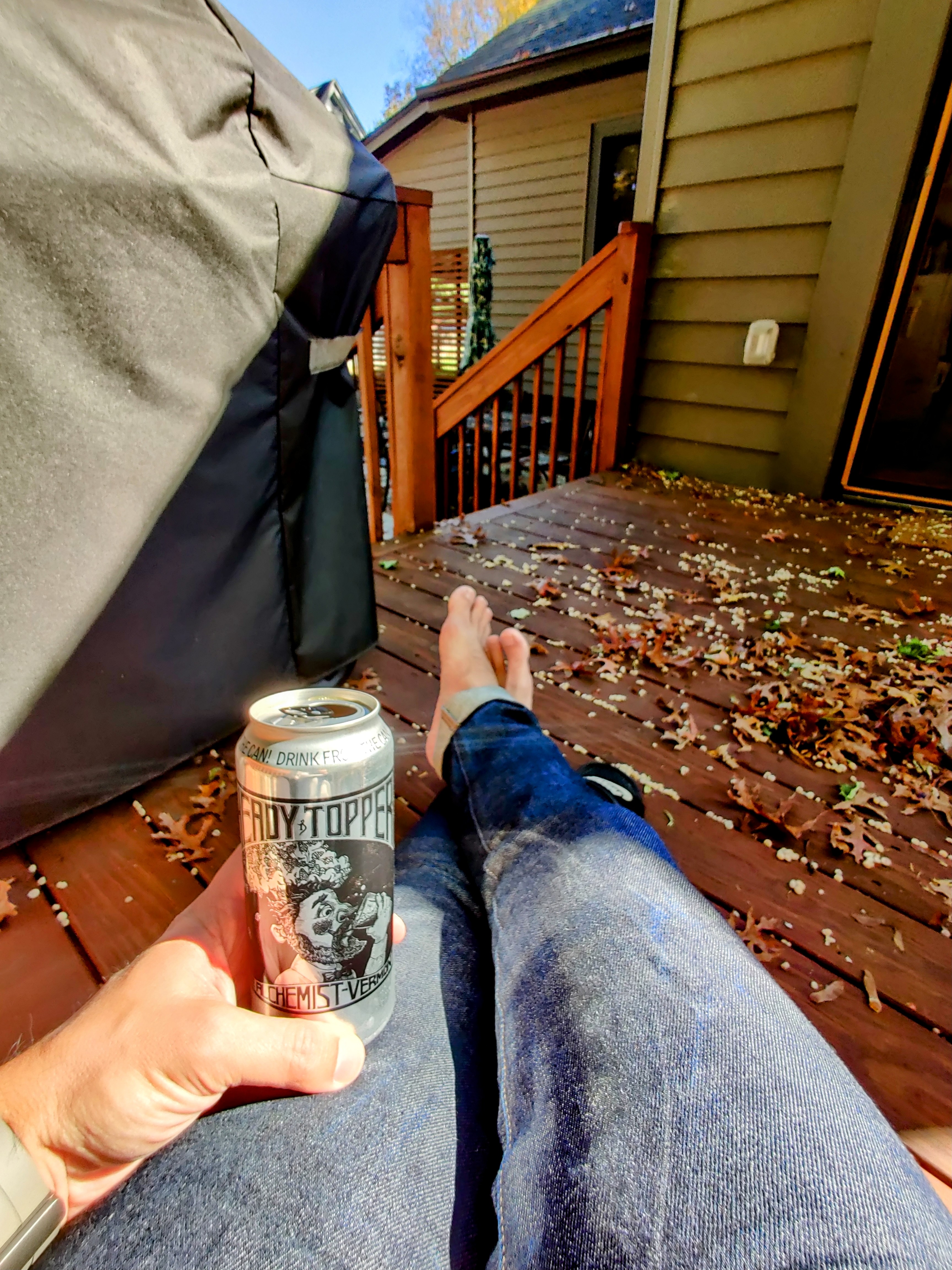 POV: enjoying some Heady Topper beer from its signature monochrome silver tallboy can, sitting in a low chair, wearing blue jeans, bare feet crossed, on a wooden deck covered in Autumn oak tree detritus.