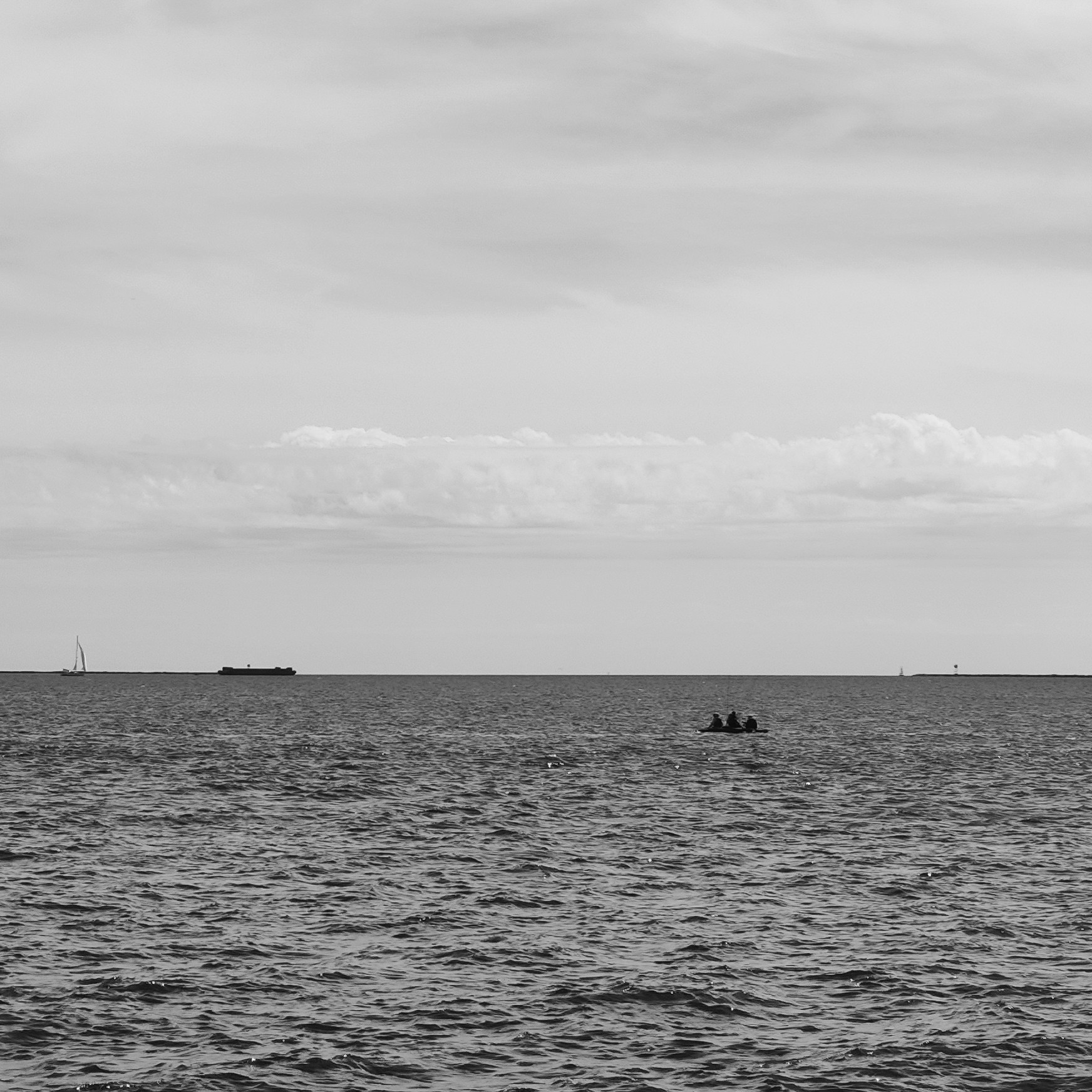 Black and white photo of New Haven Harbor from the West Haven store, showing a small boat with three fishermen in the middle ground and a barge and a sailboat on the horizon.