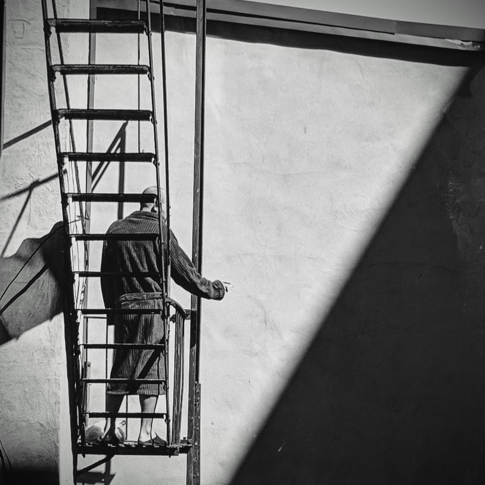 A black and white version of a photo I previously posted of a man standing on a fire escape with his back to the camera, smoking a cigarette.