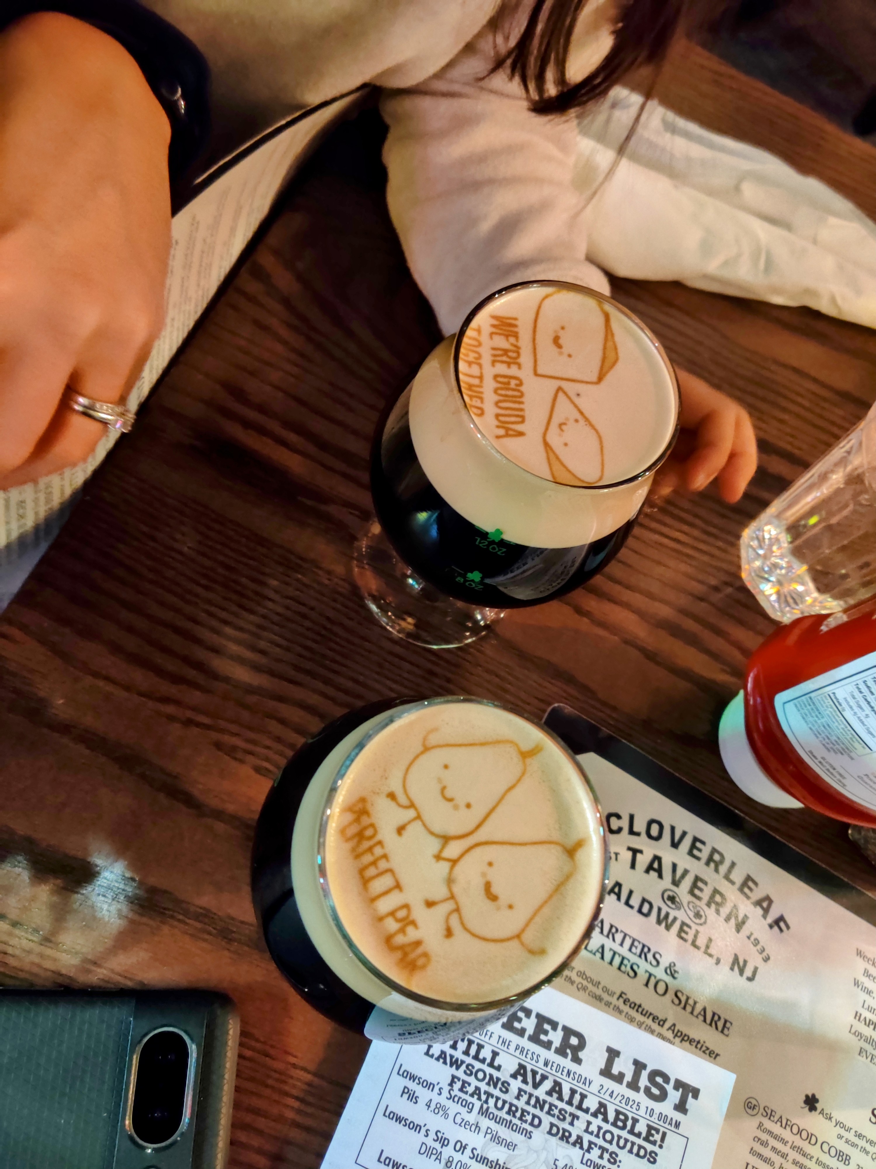 Two foamy stouts, with images printed on the foam, on a wooden tabletop. In one glass the image is of two wedges of cheese with the caption "we're Gouda together". In the other it's two pears with the caption "perfect pear".